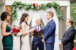 Bride in wedding dress and groom in blue suit under Ceremony wow floral design of greenery and red, coral and white blooms designed by durham florist poppy belle floral design