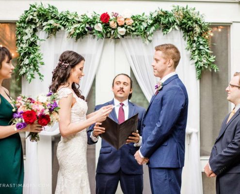Bride in wedding dress and groom in blue suit under Ceremony wow floral design of greenery and red, coral and white blooms designed by durham florist poppy belle floral design