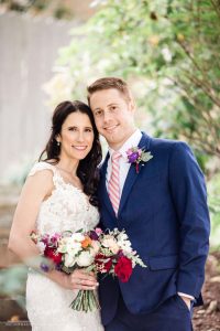 groom in blue suit next to Bride in sweetheart dress holding Red, coral, orange and white blooms with greenery bridal bouquet designed by durham florist poppy belle floral design
