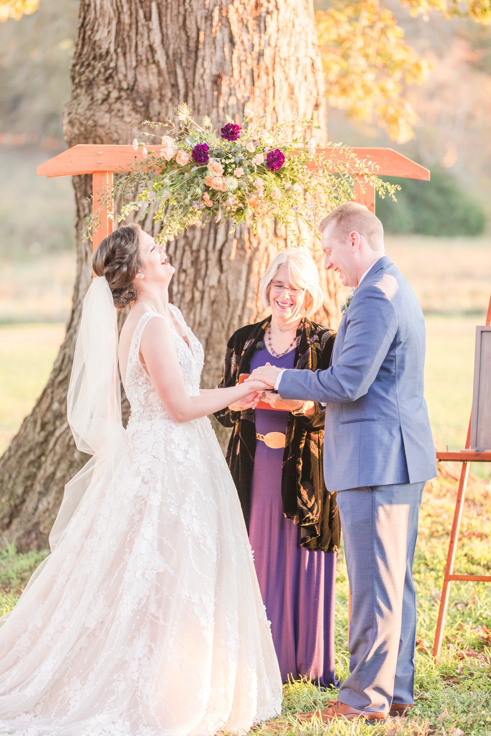 Bride in v neck lace wedding dress laughs while holding groom with blue suit under Purple, coral and white blooms with greenery pulled together in a beautiful wedding ceremony floral design created by durham florist poppy belle floral design
